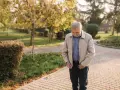 Starszy mężczyzna w beżowej kurtce Harrington i niebieskiej koszuli spaceruje po parku jesienią.