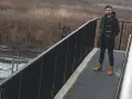Mężczyzna w budrysówce i traperach stoi na moście z widokiem na zarośla.