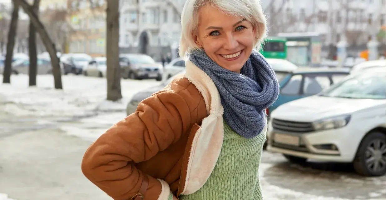 Uśmiechnięta kobieta w brązowym, sztucznym kożuszku damskim i niebieskim szaliku pozuje na tle zimowego miasta.