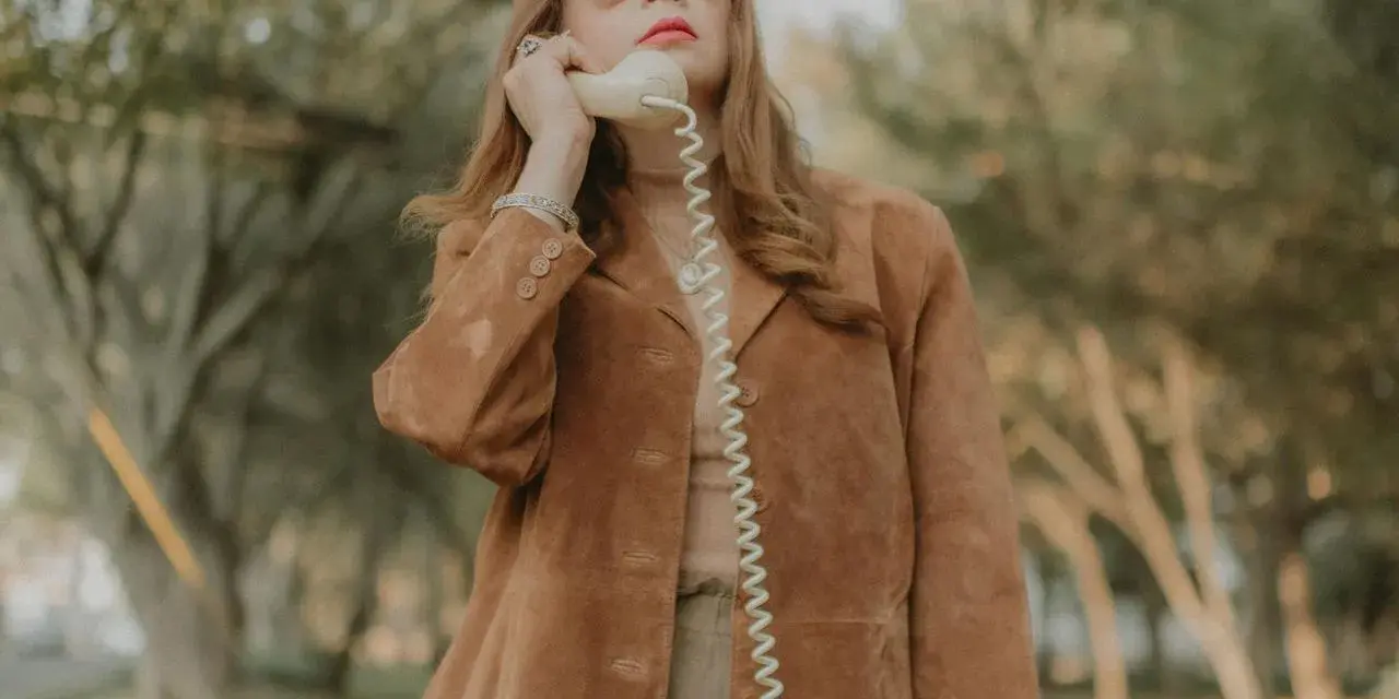 Kobieta w brązowej zamszowej kurtce damskiej rozmawia przez telefon w stylu retro.