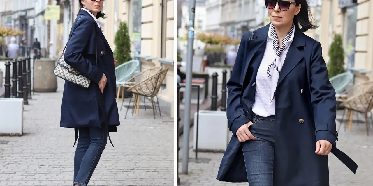 Elegancka kobieta w granatowym trenczu damskim, białej bluzce w paski i jeansach spaceruje po brukowanej ulicy.