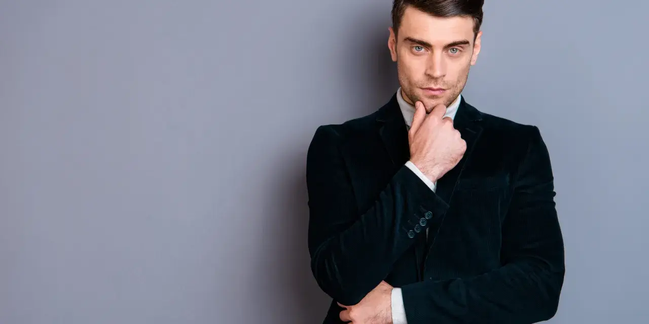 Elegancki mężczyzna w ciemnej, **marynarce aksamitnej męskiej**, z dłonią na podbródku, spogląda wprost.