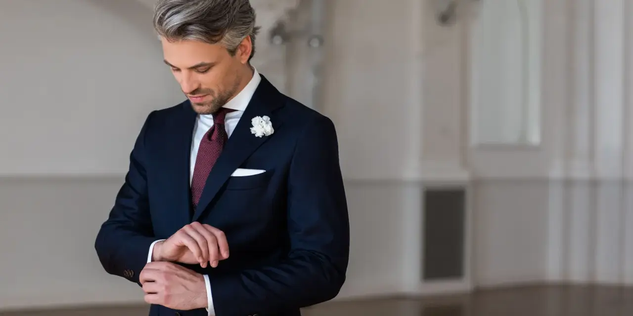 Elegancki mężczyzna w granatowym garniturze i bordowym krawacie poprawia mankiet.
