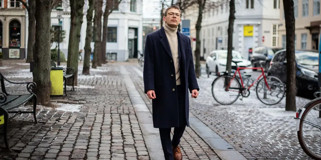 Elegancki mężczyzna w granatowym płaszczu damskim i golfie spaceruje po brukowanej ulicy. Stylowe granatowy płaszcz damski stylizacje.