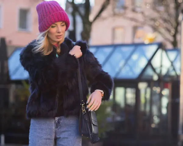 Krótkie futerko stylizacje: blondynka w różowej czapce i czarnym futerku, z jeansami i torebką.