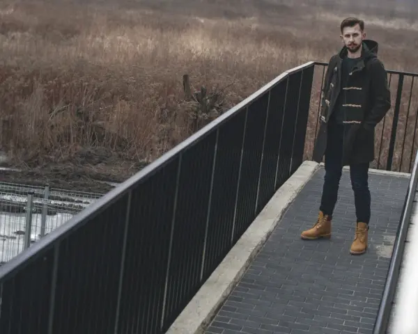Mężczyzna w budrysówce i traperach stoi na moście z widokiem na zarośla.