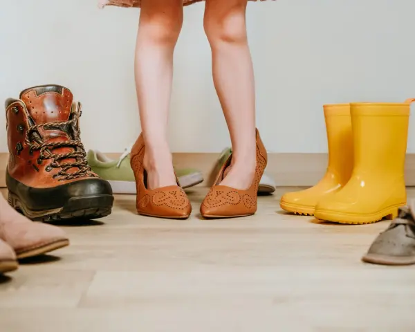 Dziecko przymierza za duże buty, szukając sposobu, jak zmniejszyć buty. Wokół różne rodzaje obuwia: botki, trapery, kalosze.