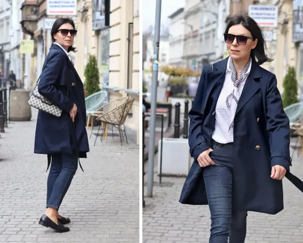 Elegancka kobieta w granatowym trenczu damskim, białej bluzce w paski i jeansach spaceruje po brukowanej ulicy.