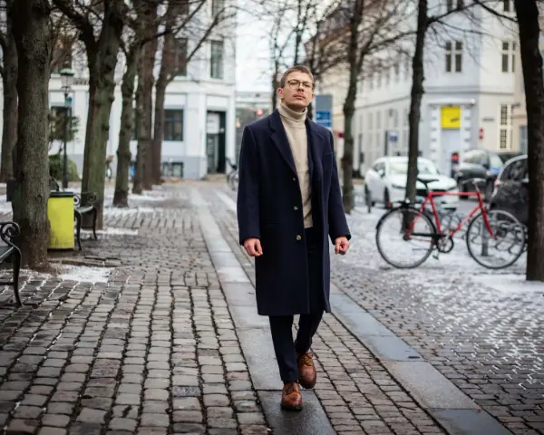Elegancki mężczyzna w granatowym płaszczu damskim i golfie spaceruje po brukowanej ulicy. Stylowe granatowy płaszcz damski stylizacje.