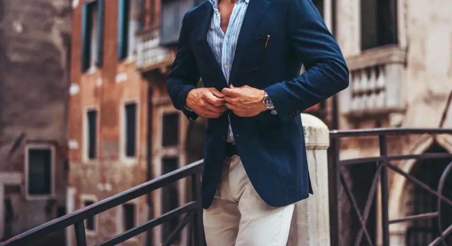 Elegancki mężczyzna w granatowej marynarce i beżowych spodniach, uosabiający włoski styl męski, zapina marynarkę na tle weneckiej kamienicy.