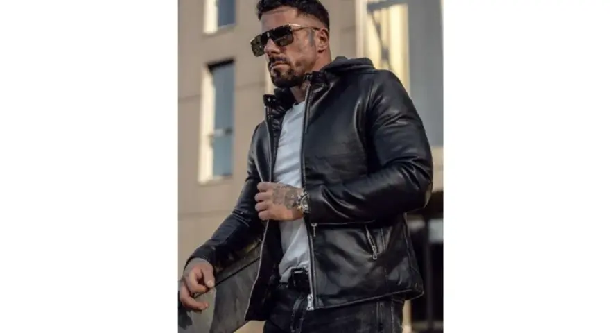 Mężczyzna w stylowej, czarnej kurtce woskowanej męskiej, z kapturem, okularach przeciwsłonecznych i tatuażami.