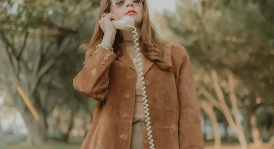 Kobieta w brązowej zamszowej kurtce damskiej rozmawia przez telefon w stylu retro.