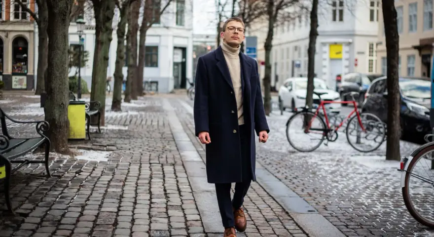 Elegancki mężczyzna w granatowym płaszczu damskim i golfie spaceruje po brukowanej ulicy. Stylowe granatowy płaszcz damski stylizacje.