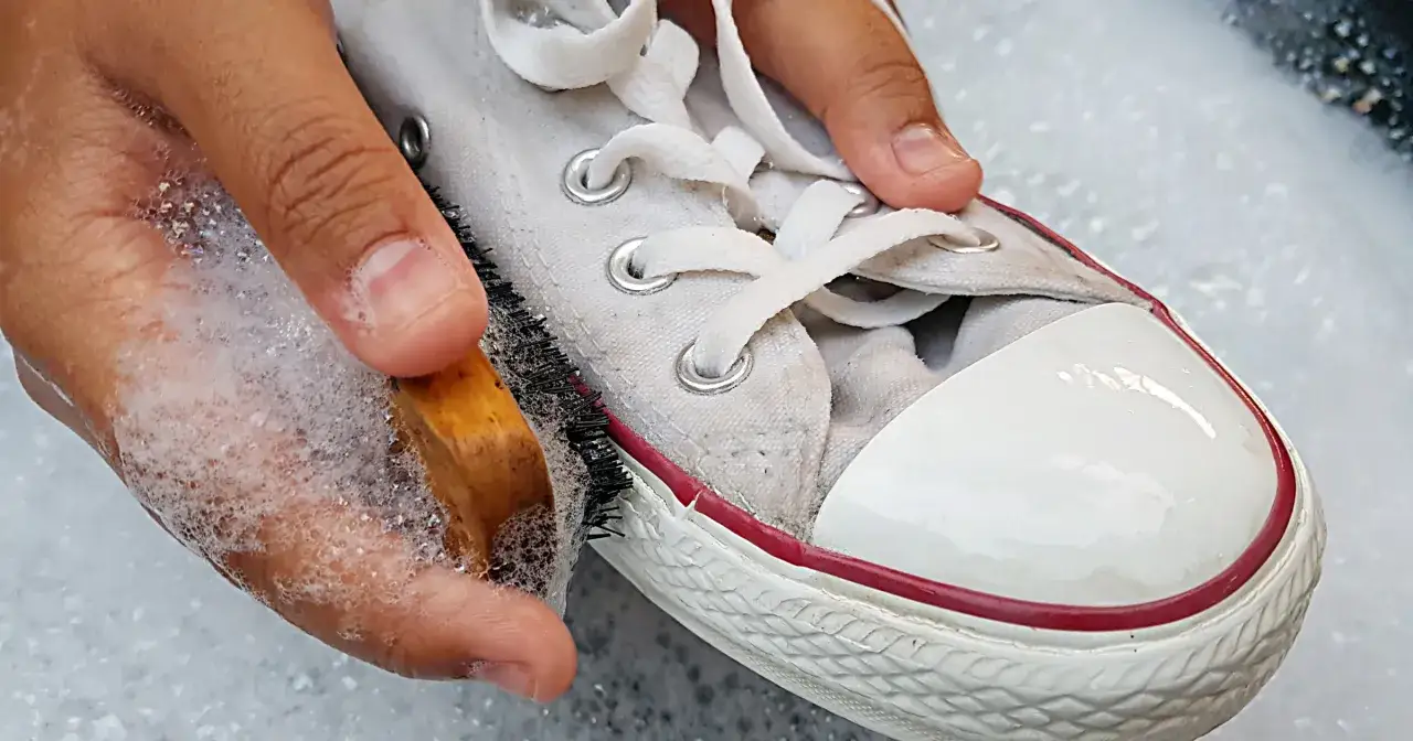 Dłoń z gąbką i szczotką czyści białe trampki w pianie. Soda do butów pomoże przywrócić im blask.