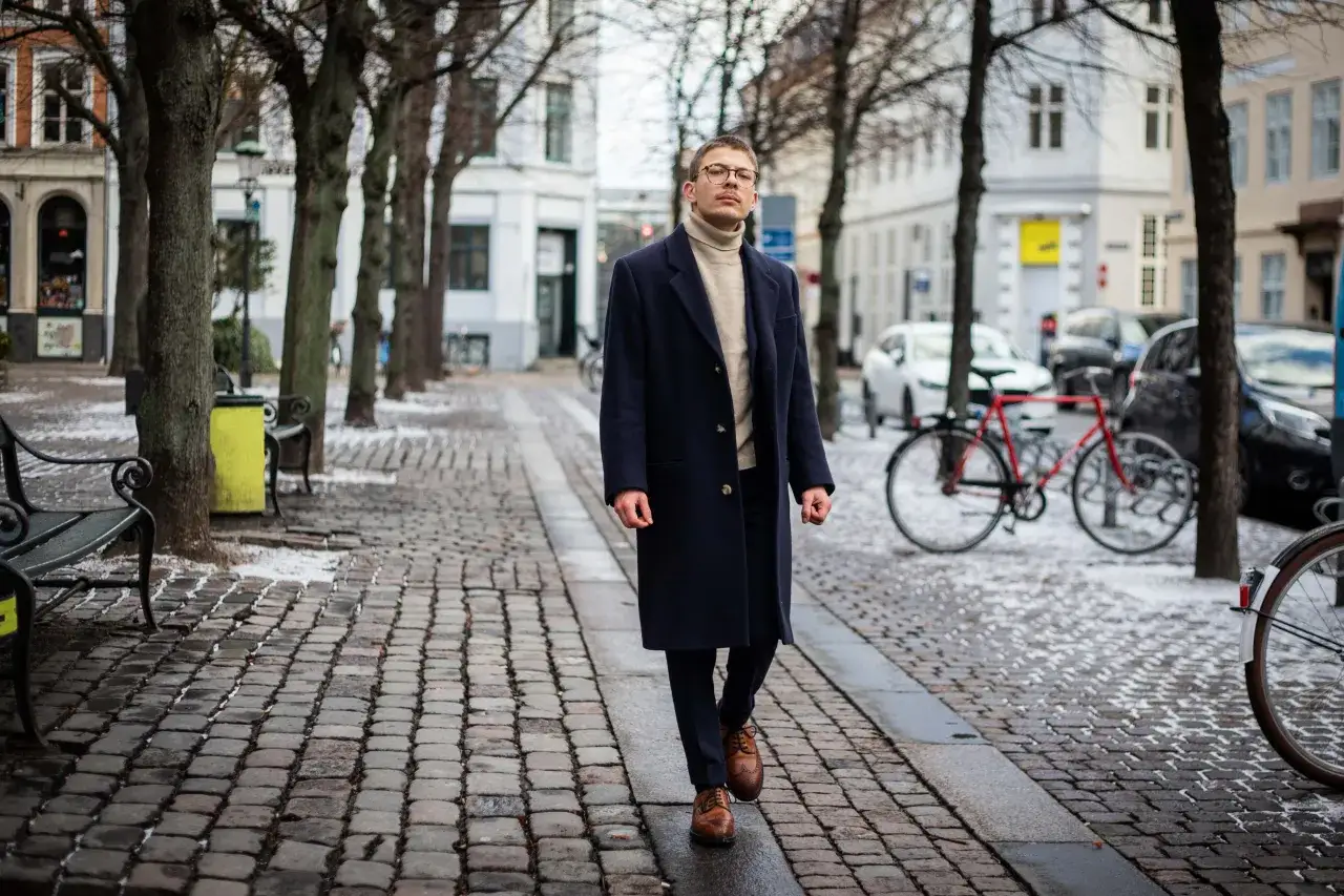 Elegancki mężczyzna w granatowym płaszczu damskim i golfie spaceruje po brukowanej ulicy. Stylowe granatowy płaszcz damski stylizacje.