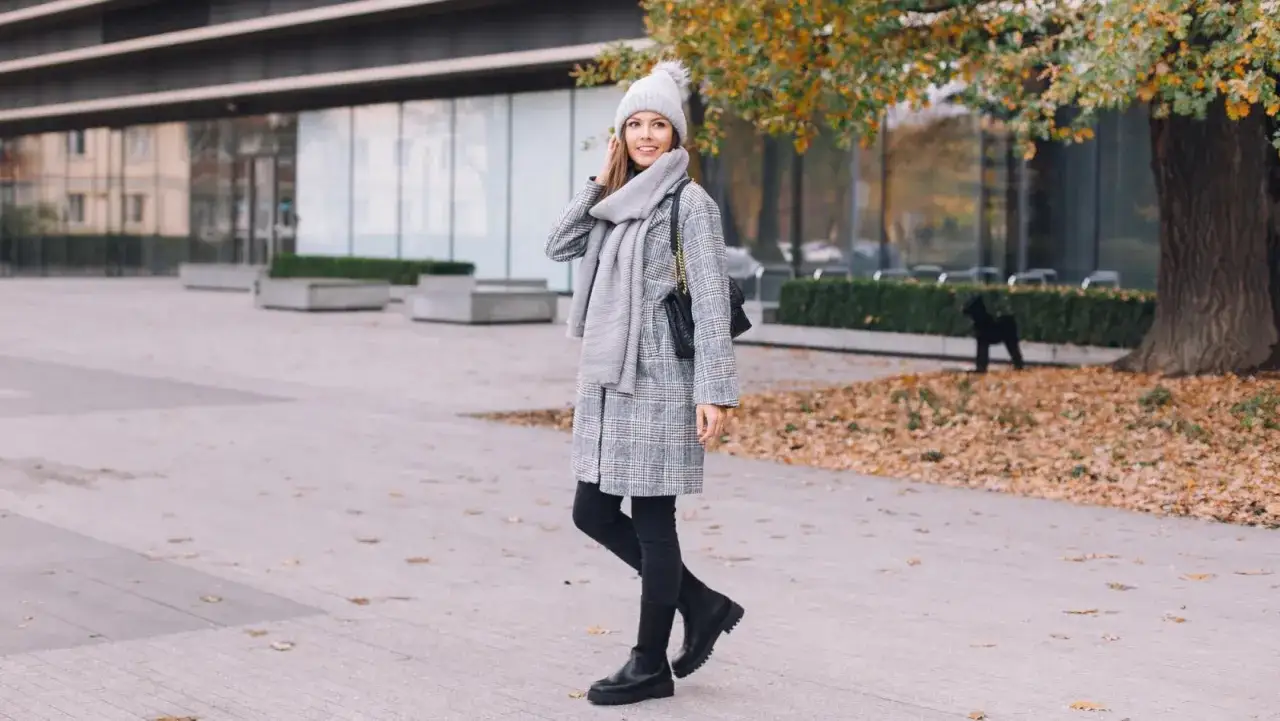 Uśmiechnięta kobieta w czapce i szaliku prezentuje modną zimową stylizację damską: kraciasty płaszcz, czarne rurki i botki.