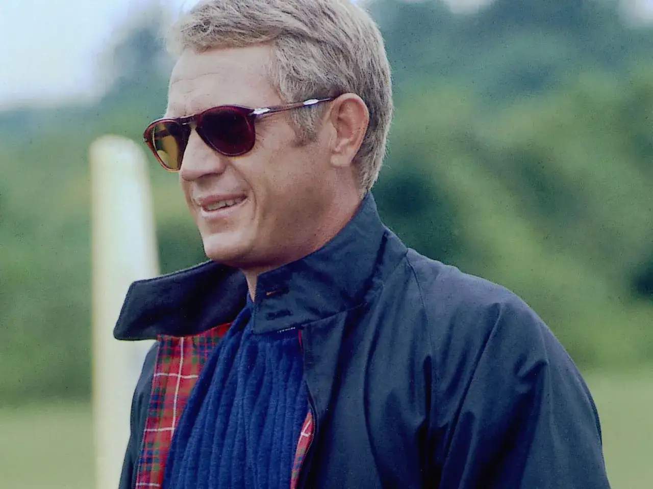 Steve McQueen w granatowej kurtce typu harrington z podszewką w kratę, niebieskim swetrze i okularach przeciwsłonecznych.