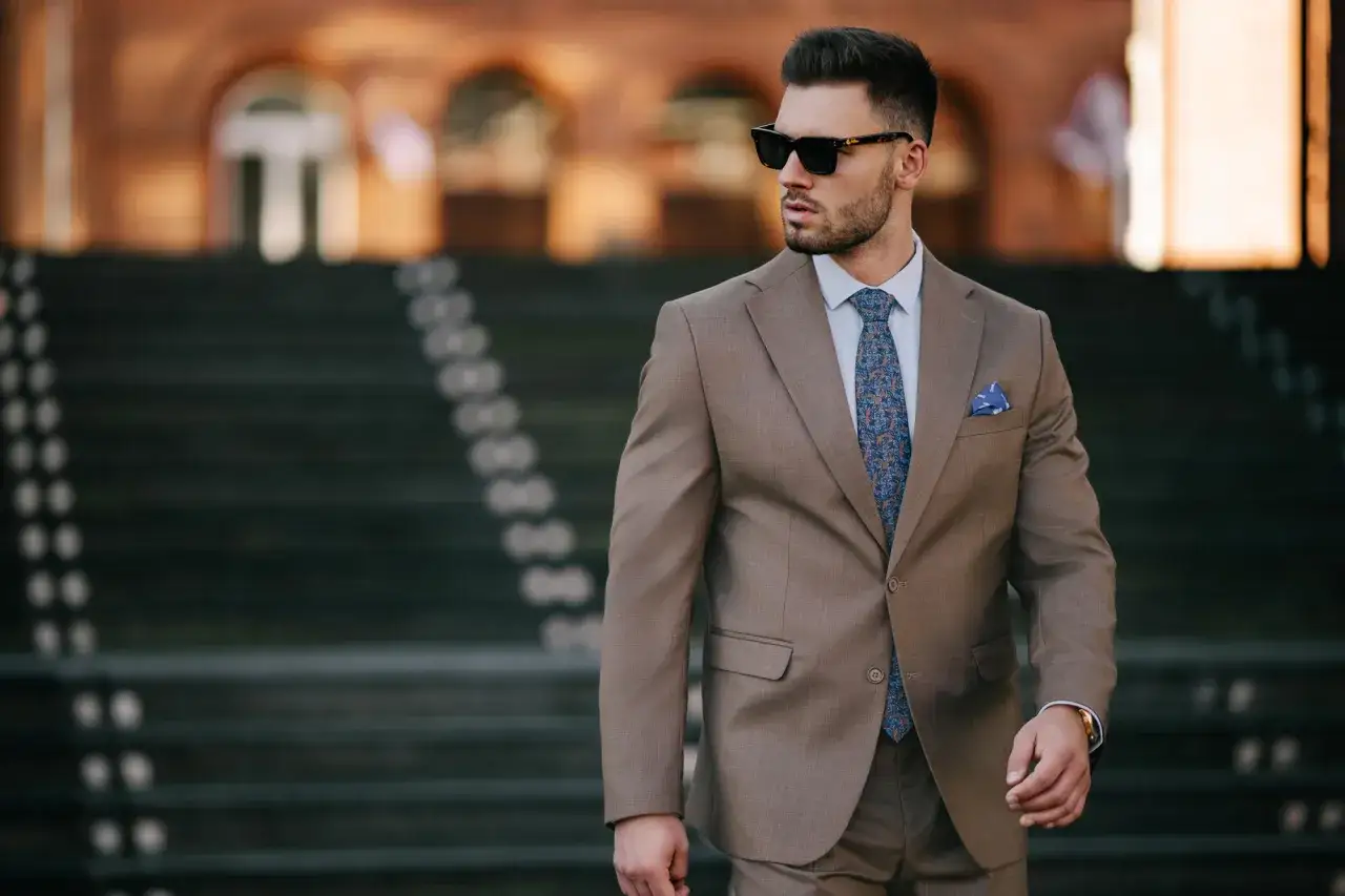 Elegancki mężczyzna w brązowym garniturze, z niebieską poszetką i wzorzystym krawatem. Idealny przykład, jak dobrać poszetkę do stylizacji.