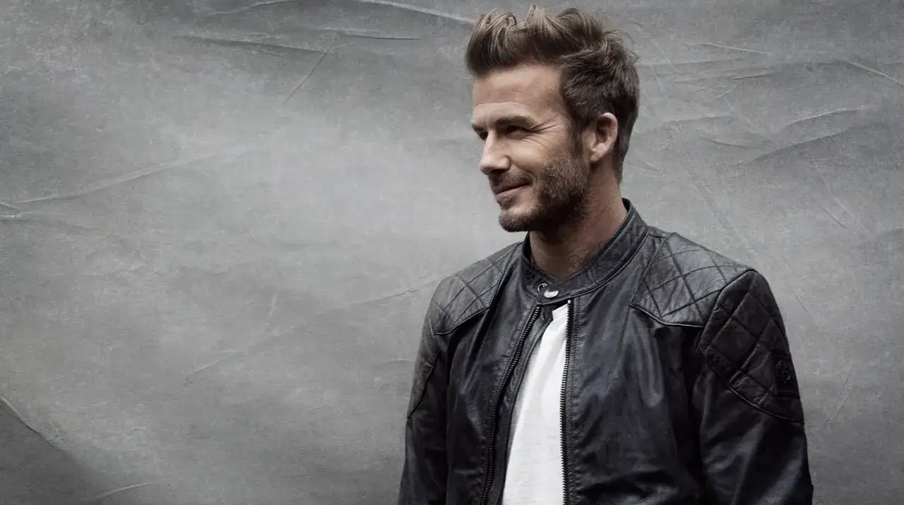 David Beckham w stylowej, pikowanej kurtce woskowanej męskiej, z uśmiechem spogląda w bok na tle szarej ściany.