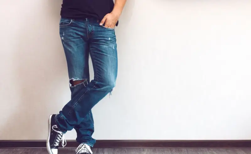 Mężczyzna w modnych jeansach z dziurami na kolanie, czarnych trampkach i czarnej koszulce, opiera się o ścianę.